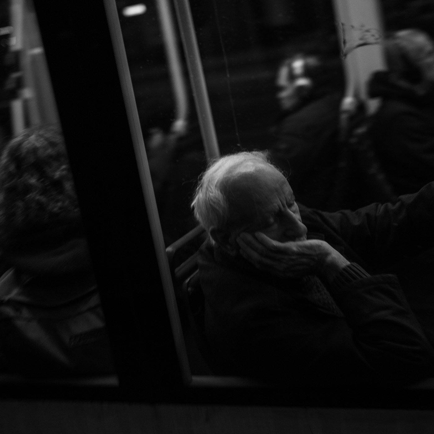 An elderly man, seated on a tram at night, his face resting in his hand