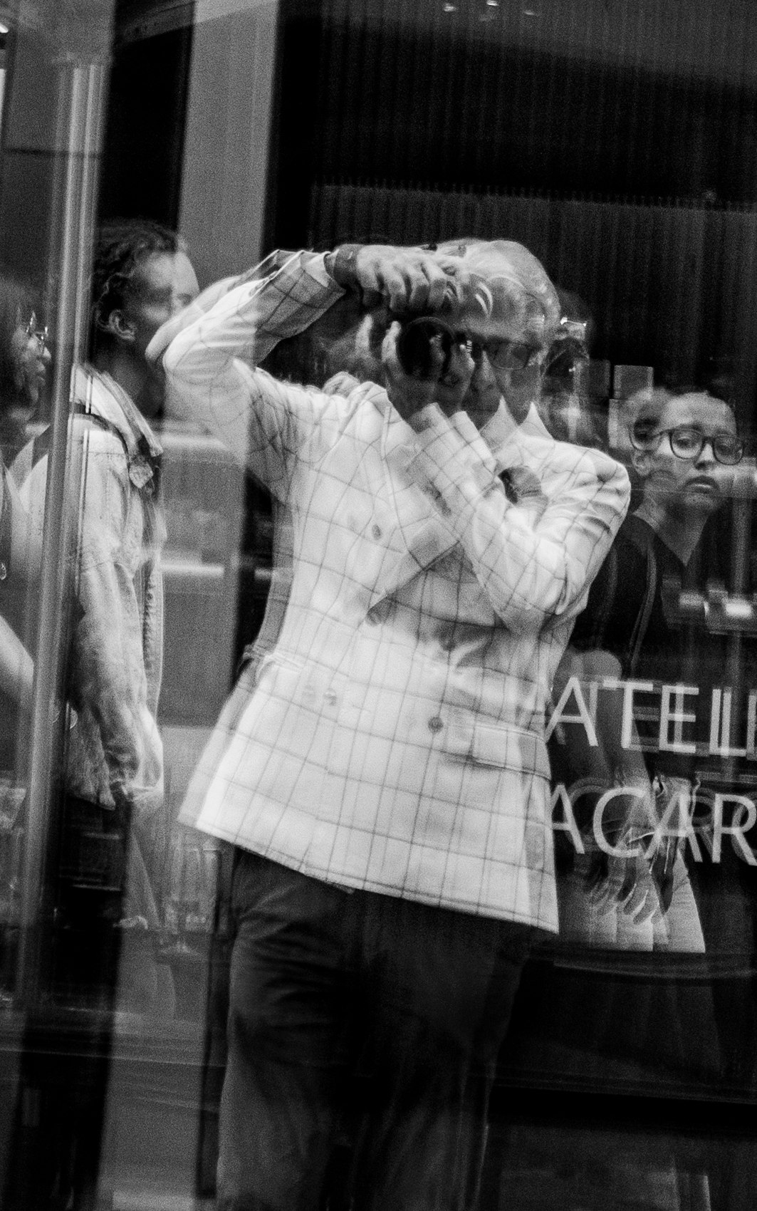 Le photographe se reflète dans une vitrine, entouré de passants et de reflets de la rue