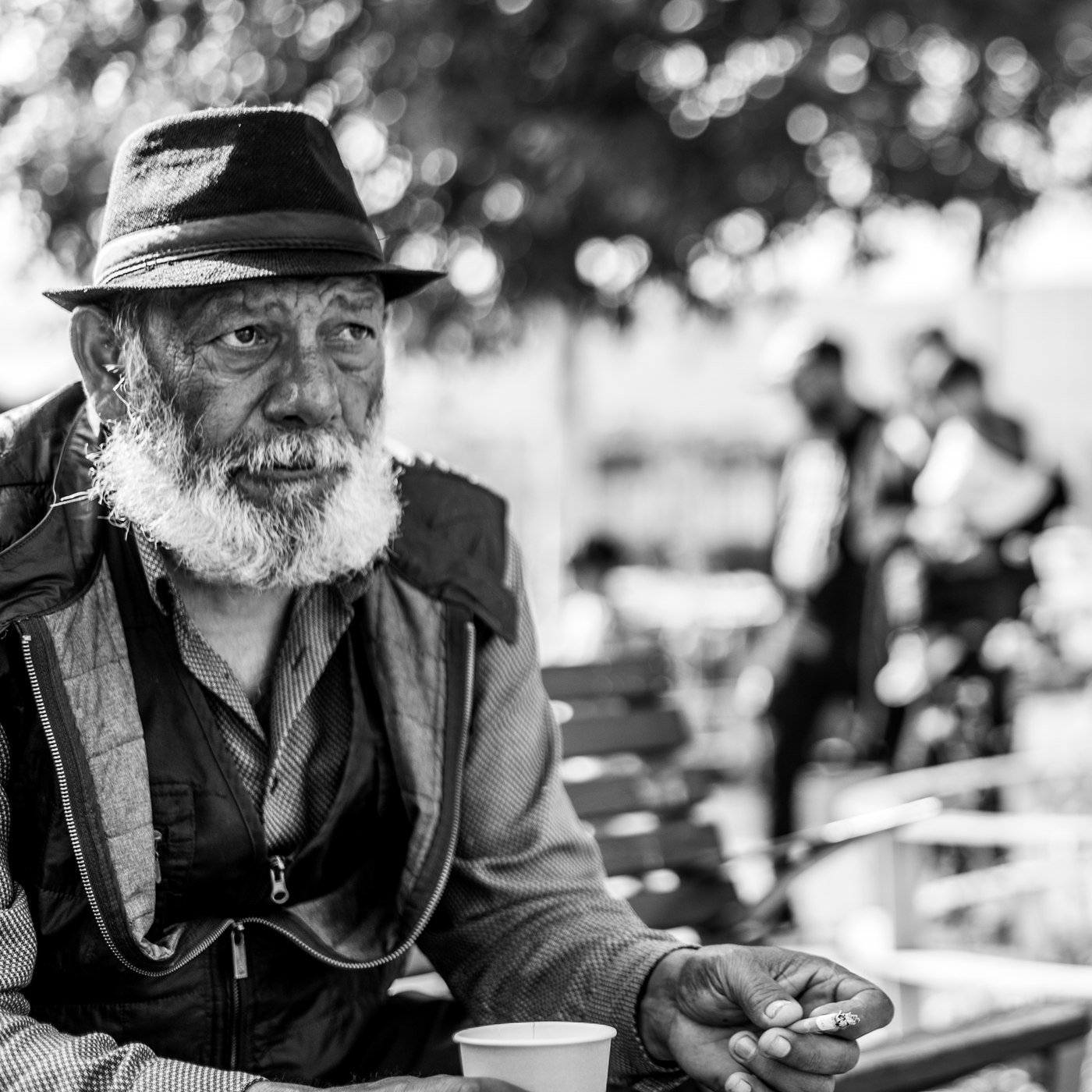 Vasile, assis sur un banc à Plainpalais, chapeau et barbe blanche, une cigarette à la main